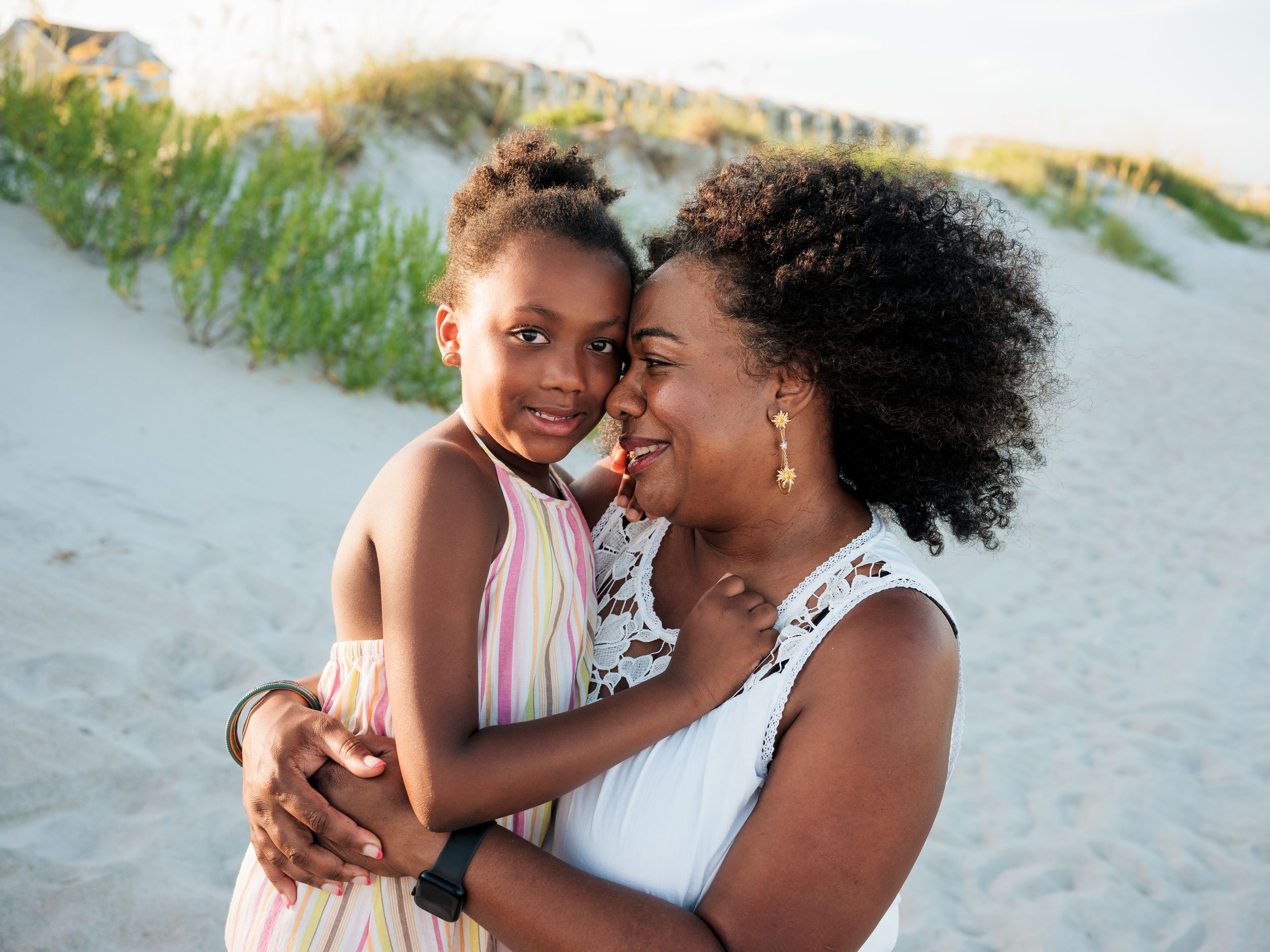 A woman and young girl share a tender moment on a sunny beach, touching foreheads and smiling.