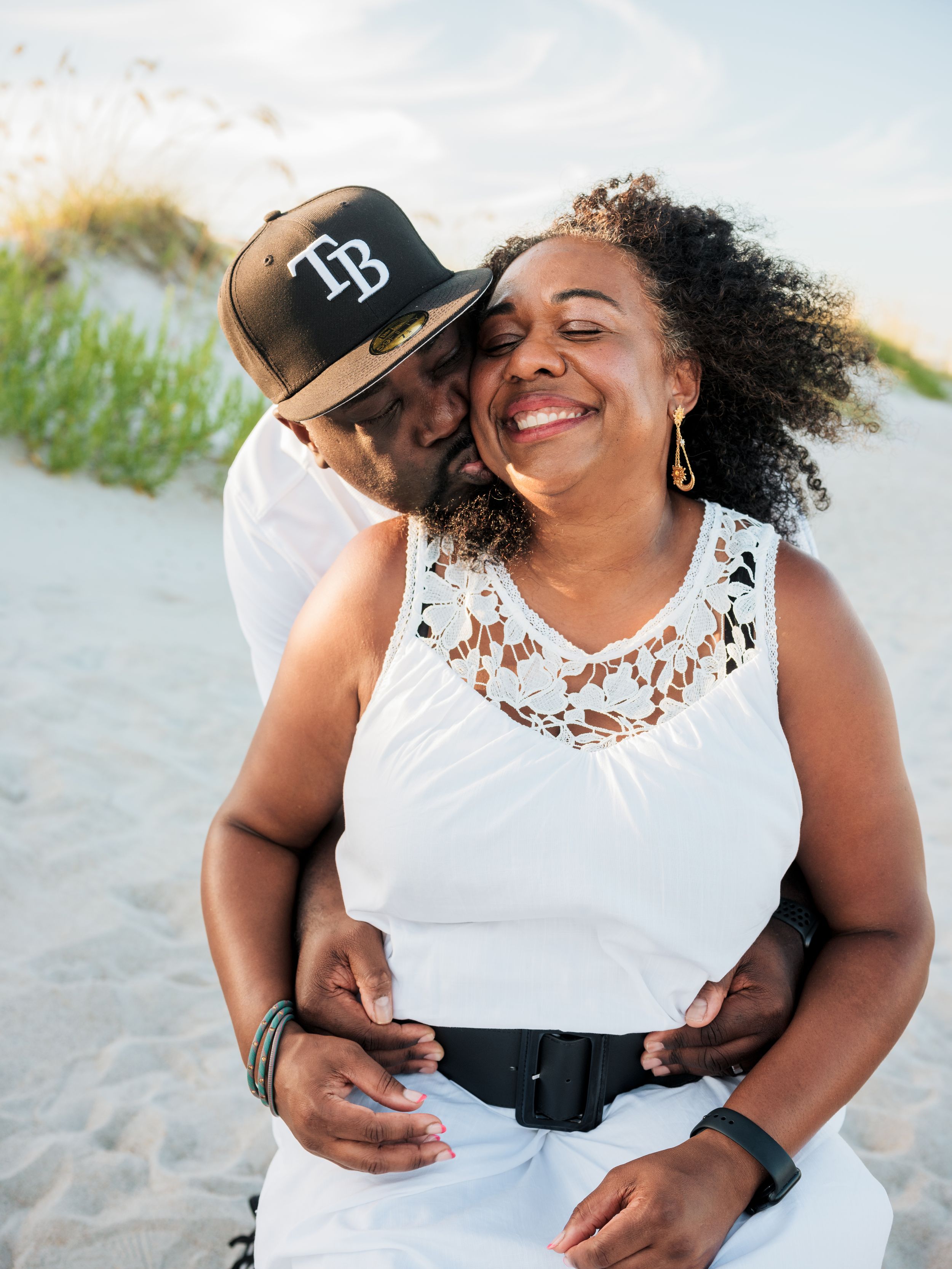 A joyful couple embraces on a sandy beach, the man kissing her cheek while she smiles, both wearing white outfits.