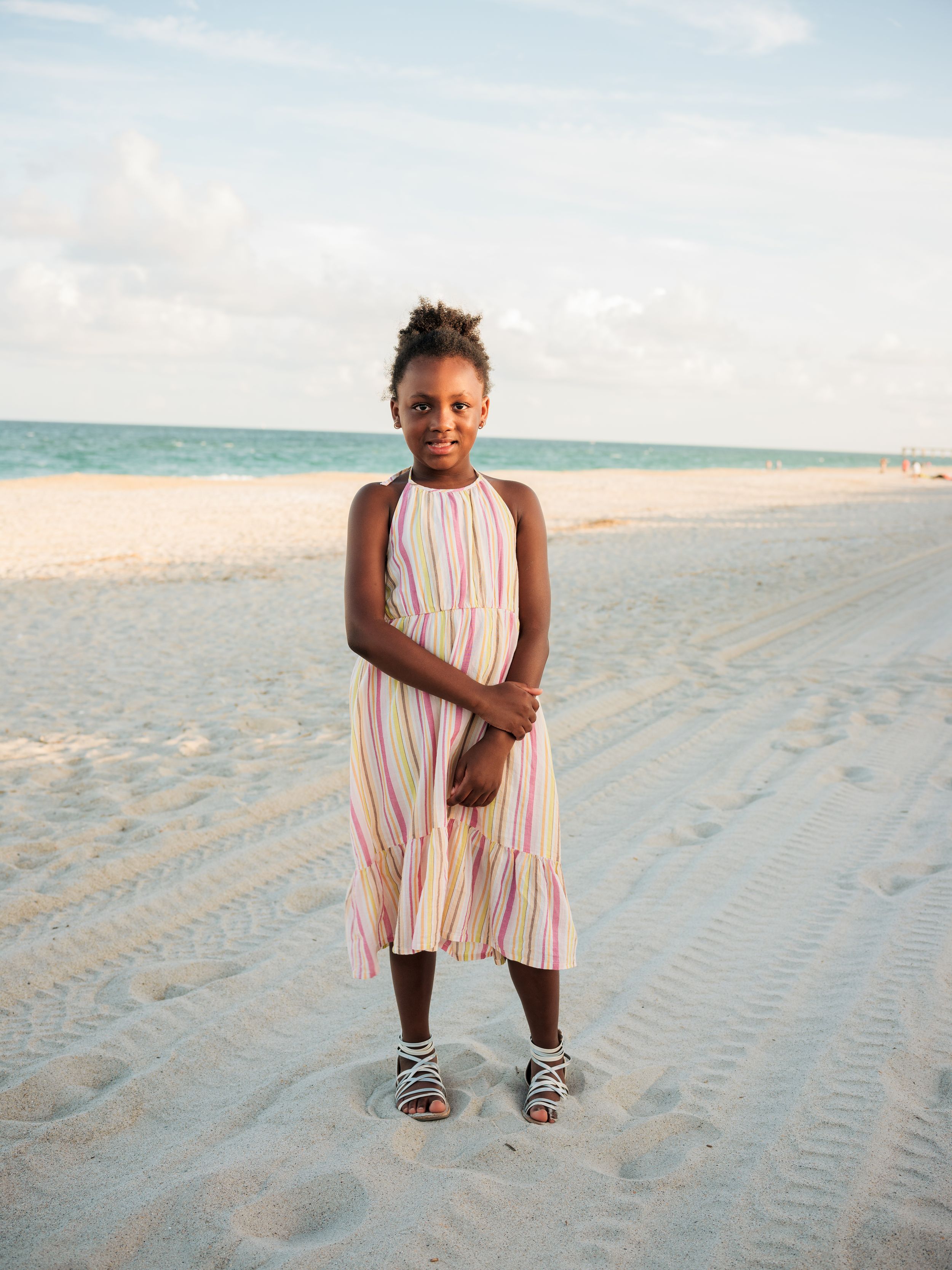 Young girl in striped dress standing on sandy beach with turquoise ocean and cloudy sky behind her.