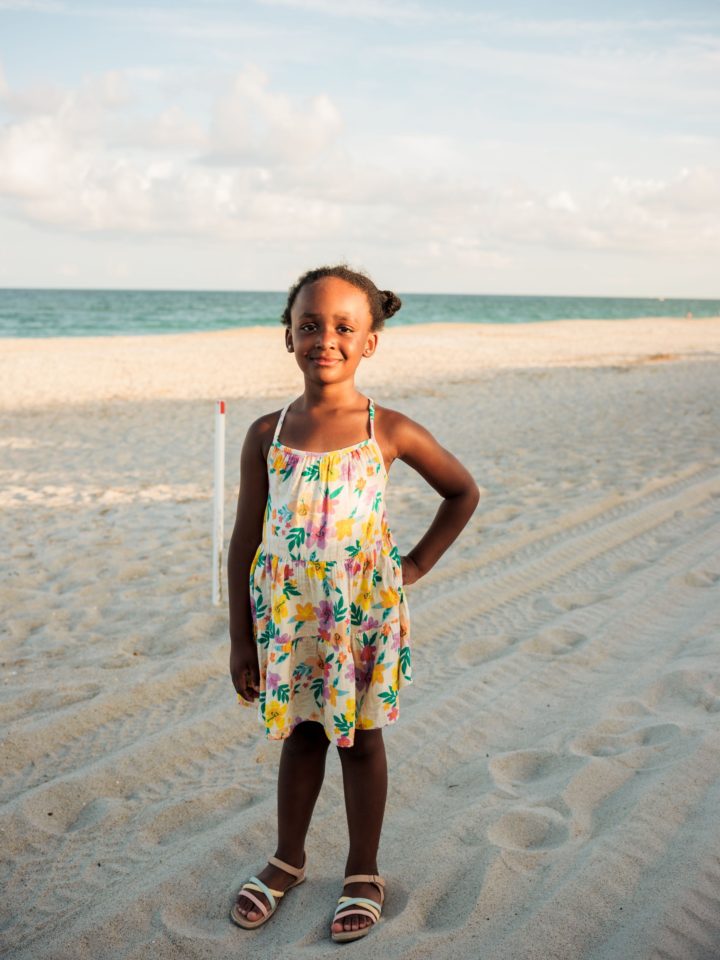 Young girl in floral sundress smiling on sandy beach with ocean horizon backdrop.