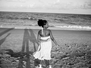Black and white photo of a woman in a white dress walking on a sandy beach toward ocean waves.