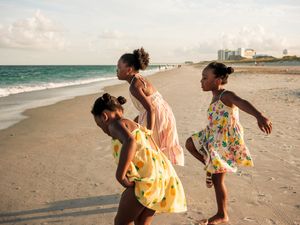 Three young girls in colorful dresses playing on a sandy beach with ocean waves and city skyline in background.