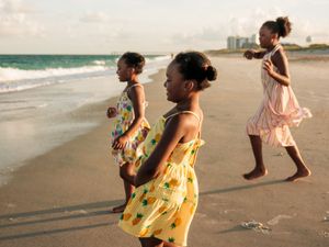 Three young girls in colorful dresses playing on a sandy beach with ocean waves in the background.