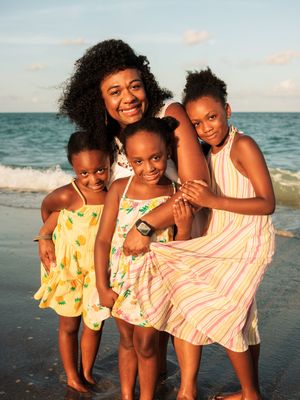 A smiling mother poses with her three young daughters on a sunny beach, all wearing colorful summer dresses.