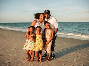 Happy family of five smiling together on a sunny beach with turquoise ocean waves in the background.