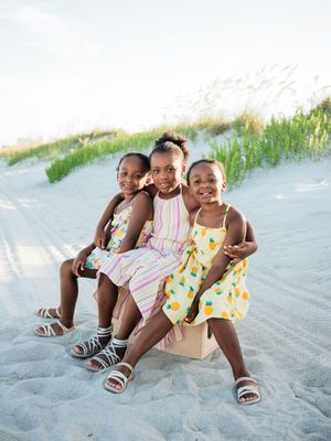 Three young girls in colorful summer dresses sit together smiling on a sandy beach with green dunes behind them.
