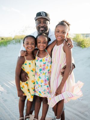 A man wearing a TB cap poses with three young girls on a sandy beach, all smiling in colorful summer dresses.