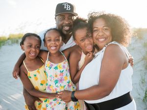 Happy Black family of five smiling together on a sandy beach at sunset, parents with three young daughters.