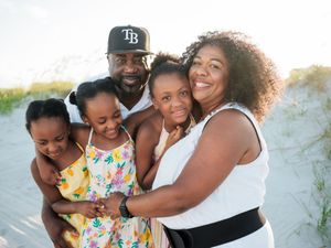 Happy Black family of five smiling together on a sandy beach, parents with three daughters in summer outfits.