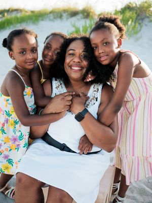 A smiling mother sits on a beach surrounded by her three young daughters hugging her warmly on a sunny day.