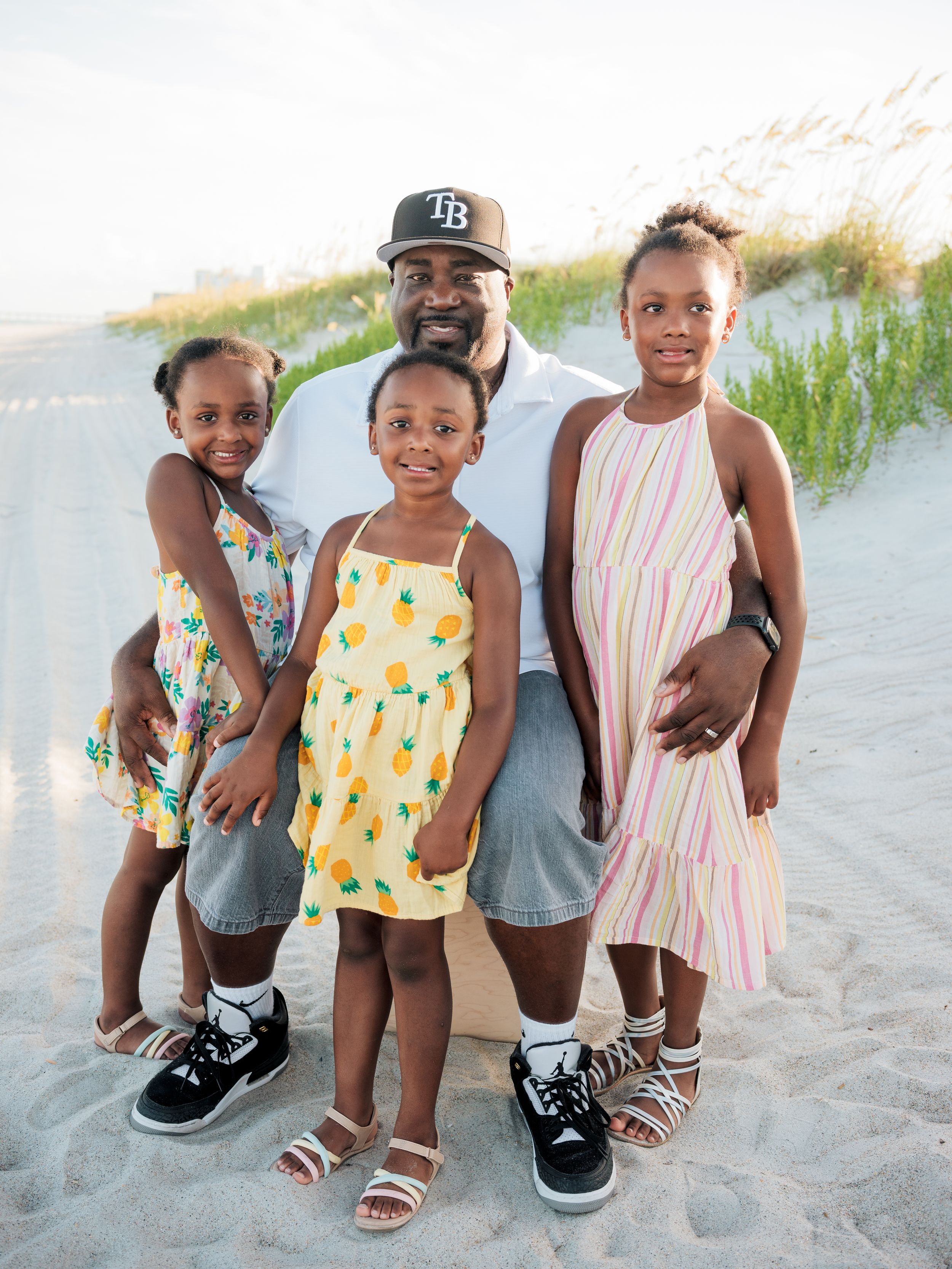 A man and three young girls pose together on a sandy beach, smiling in colorful summer outfits.