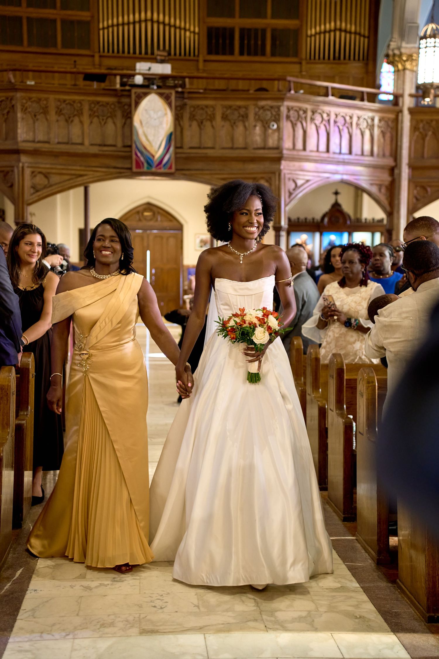 Elegant Black bride in a strapless white gown with veil holds a bouquet while walking down a church aisle.