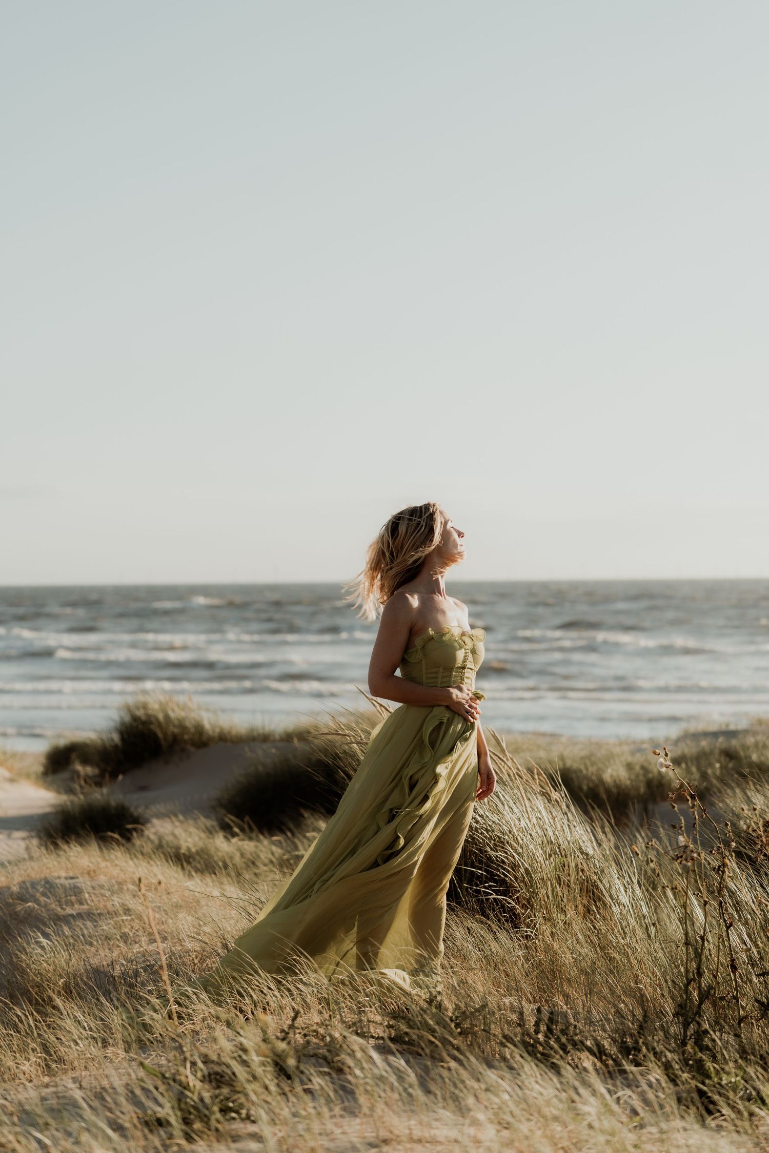 Woman in flowing yellow dress standing on coastal dunes with ocean waves in the background.