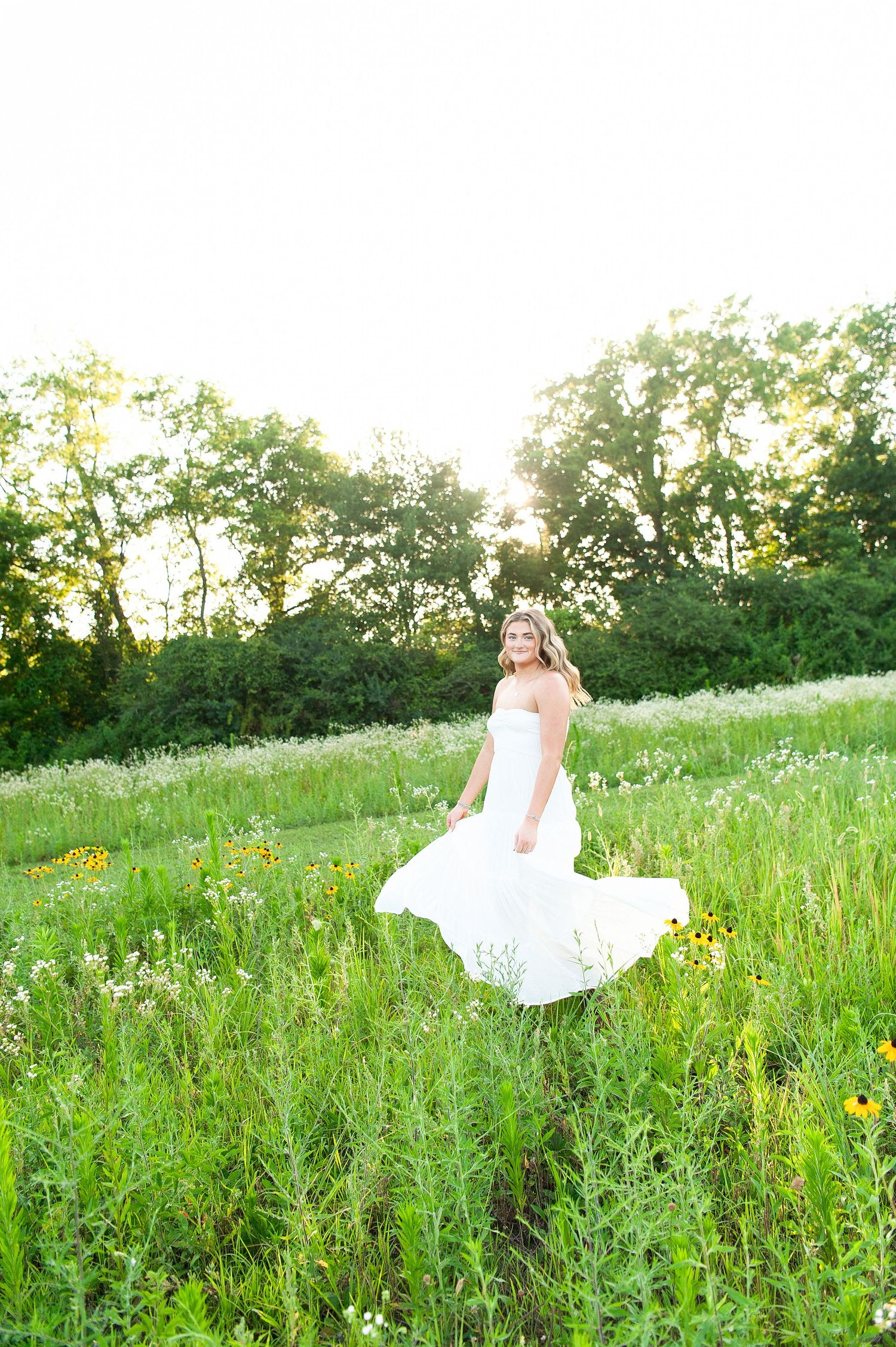 Bride in white gown twirling in a sunlit wildflower meadow with lush green trees in the background.