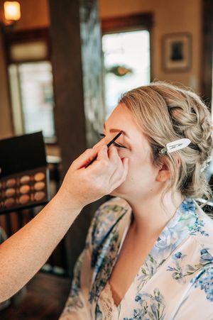 Makeup artist applying eye makeup to a blonde bride in a floral robe, with a makeup palette visible in the background.