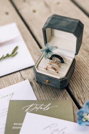 Elegant wedding rings displayed in a gray velvet box surrounded by wedding stationery on a rustic wood surface.