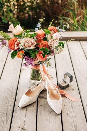 Pearl bridal flats with ribbon ties beside a vibrant wedding bouquet of orange, pink, and purple blooms on a wooden deck.