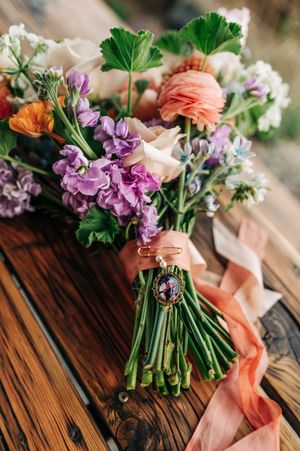 Colorful wedding bouquet with purple, peach, and orange flowers, green stems tied with a ribbon and vintage photo charm.
