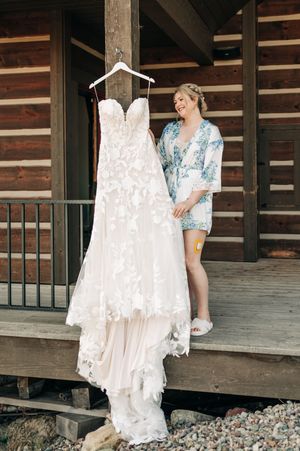 Bride in floral robe admiring her lace wedding gown hanging on a rustic log cabin porch.