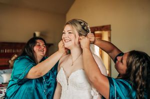 Bridesmaids in teal robes help a smiling bride put on her necklace while she gets ready in her wedding dress.