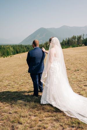 Bride and groom walk hand-in-hand through a mountain meadow, her lace gown and veil trailing behind.