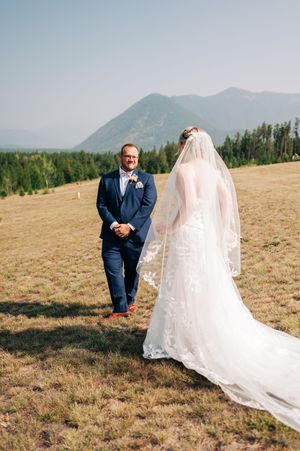 Groom in navy suit smiles at bride in lace wedding gown with cathedral veil on mountain meadow.