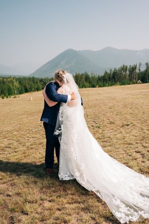 Bride and groom embrace in tender first look moment on scenic mountain meadow with forest backdrop.