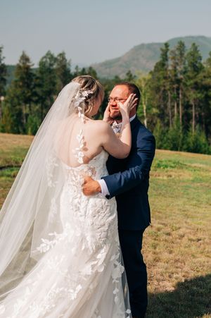 Emotional groom wiping tears as bride in lace gown and veil embraces him at outdoor mountain wedding.