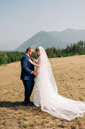 Bride and groom share a romantic moment in a mountain meadow, with the bride's lace veil flowing in the breeze.