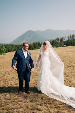 Bride and groom walk hand-in-hand in a mountain meadow, her lace gown and veil flowing in the breeze.