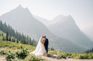 Bride and groom embrace on a mountain trail with stunning misty peaks and pine forests in the background.