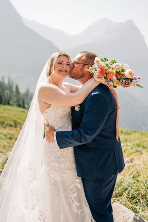 Bride smiling as groom kisses her cheek, holding a colorful bouquet with misty mountains in the background.