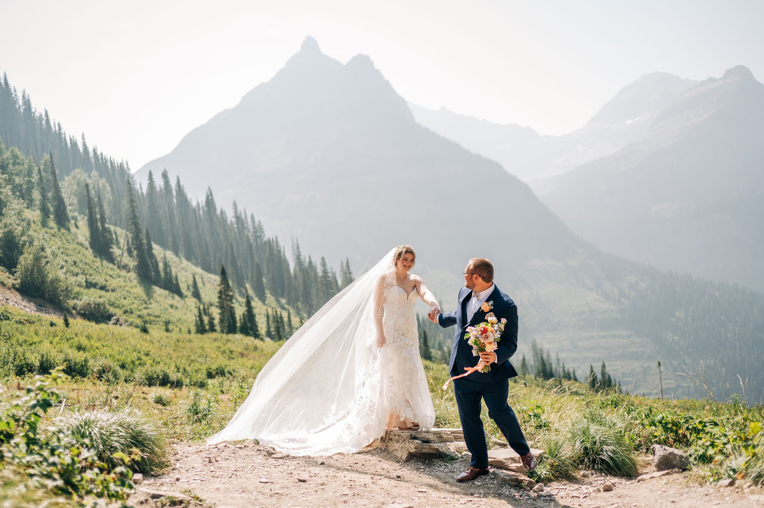 Bride and groom walk mountain trail in Glacier National Park, her veil flowing in wind, stunning peaks behind them.