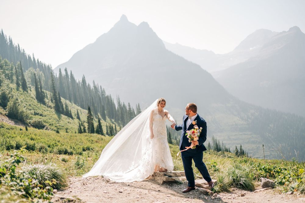 Bride and groom walk mountain trail in Glacier National Park, her veil flowing in wind, stunning peaks behind them.