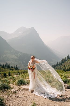Bride in lace gown with flowing veil holds colorful bouquet against stunning mountain backdrop at Glacier National Park.