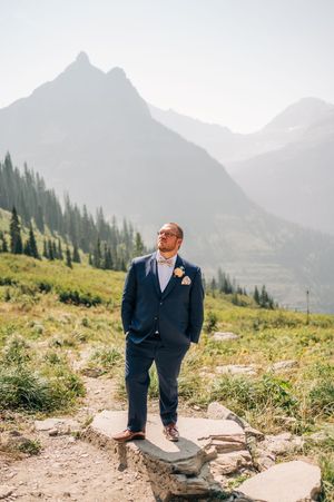 Groom in navy suit stands on rocky mountain trail with stunning peaks and evergreen forest backdrop.