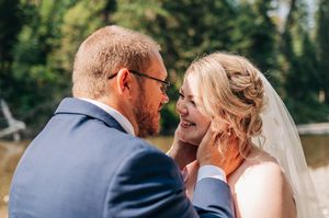 Groom tenderly cups bride's face as they share an intimate moment outdoors near a lake on their wedding day.