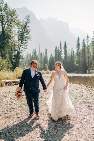 Bride and groom holding hands walking by mountain lake, bride in strapless lace gown, groom in navy suit.