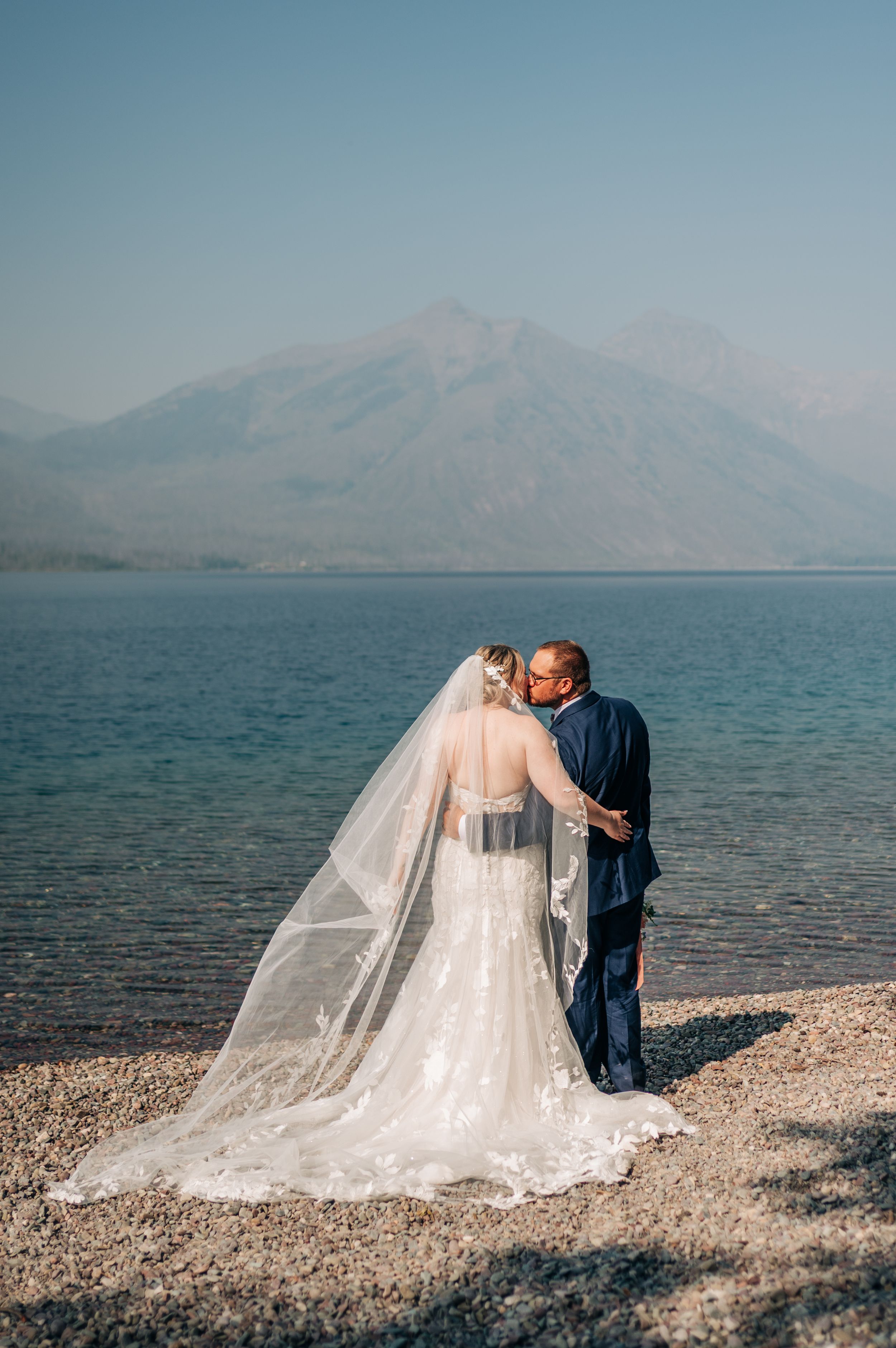 Bride and groom sharing a kiss by a serene mountain lake, her lace veil trailing on the rocky shore.