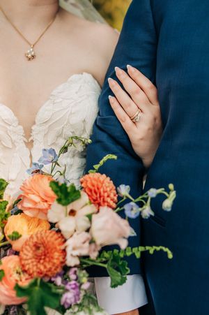 Bride holding colorful wedding bouquet with orange dahlias, wearing gold rings and lace gown beside groom in navy suit.
