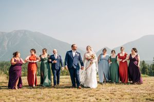 Wedding party in colorful gowns and blue suit pose with mountains backdrop on sunny day.