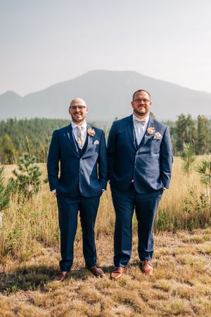 Two grooms in matching navy suits with boutonnieres stand in a mountain meadow on their wedding day.