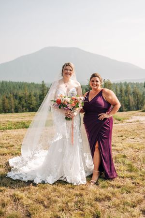 Bride in lace gown holding colorful bouquet poses with bridesmaid in purple dress against a stunning mountain backdrop.