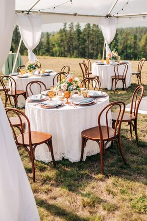 Elegant outdoor wedding reception under white tent with bentwood chairs and floral centerpieces on lace tablecloths.