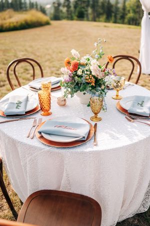 Elegant outdoor wedding reception table with white lace cloth, amber glassware, copper chargers, and colorful floral centerpiece.