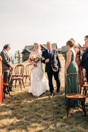 Bride in lace gown walks down outdoor aisle with escort past bentwood chairs at mountain wedding ceremony.