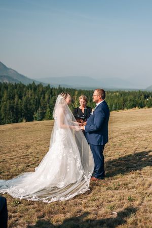 Outdoor mountain wedding ceremony with bride in lace gown and groom in navy suit exchanging vows.