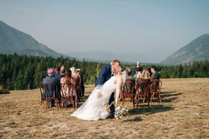 Groom dips bride for a kiss at outdoor mountain wedding ceremony with guests cheering in background.