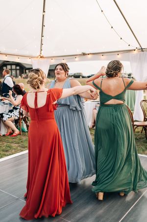 Three bridesmaids in colorful red, blue, and green gowns dancing at an outdoor wedding reception.