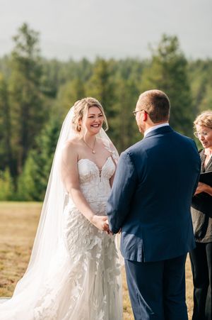 Bride in lace gown and veil laughs joyfully while holding hands with groom in navy suit during outdoor wedding ceremony.